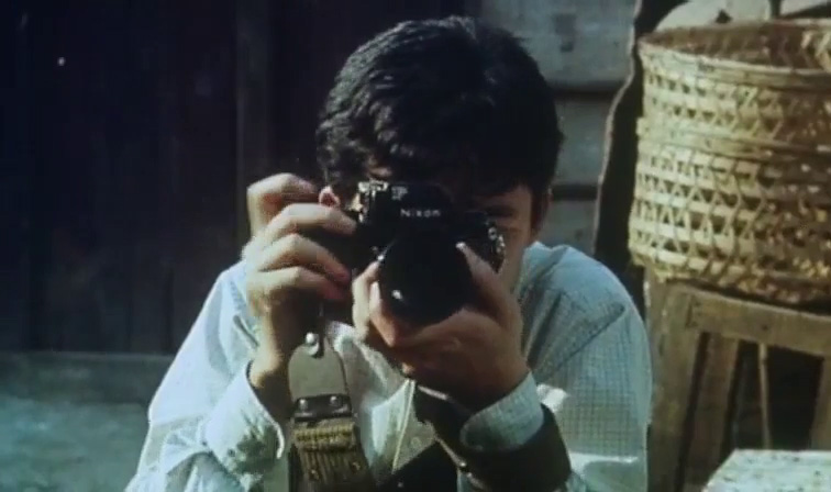 A man behind a camera squats beside some weaved baskets